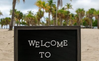 Un panneau « Bienvenue à La Havane » posé sur une plage de sable bordée de palmiers sous un ciel nuageux.